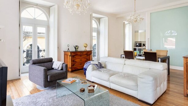 A spacious living room with a grey sofa, a white sofa, a wooden dresser, and a glass coffee table. There are two chandeliers hanging from the ceiling.