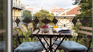 A balcony with a table, two patterned cushions, a bowl of grapes, and a book.