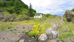 A white house on a grassy hill with a rocky foreground and a stream. - The ultimate secluded Holiday Cottage (Oban)