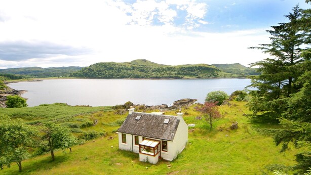 A small white house with a sloped roof, situated on a grassy hill overlooking a large body of water and distant hills. - The ultimate secluded Holiday Cottage (Oban)