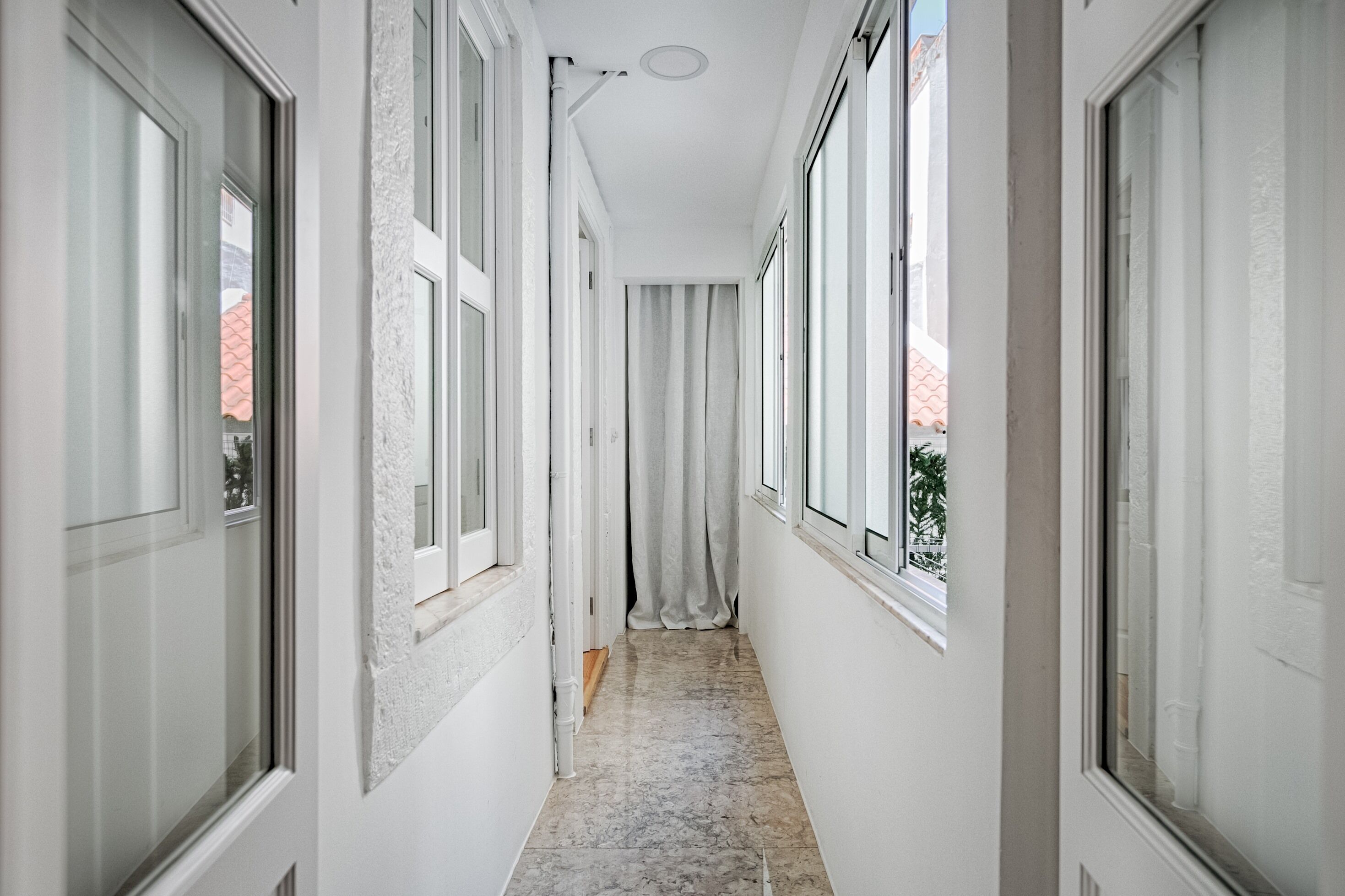 A narrow hallway with white walls and large windows on both sides.