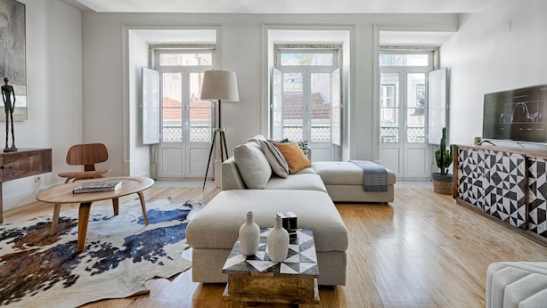 Ein geräumiges Wohnzimmer mit einer großen Couch, einem Couchtisch und einem Fernseher. Große Fenster bieten einen Blick auf die umliegenden Gebäude.