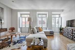 A spacious living room with a large sofa, a coffee table, and a TV. There are large windows and a view of buildings outside.