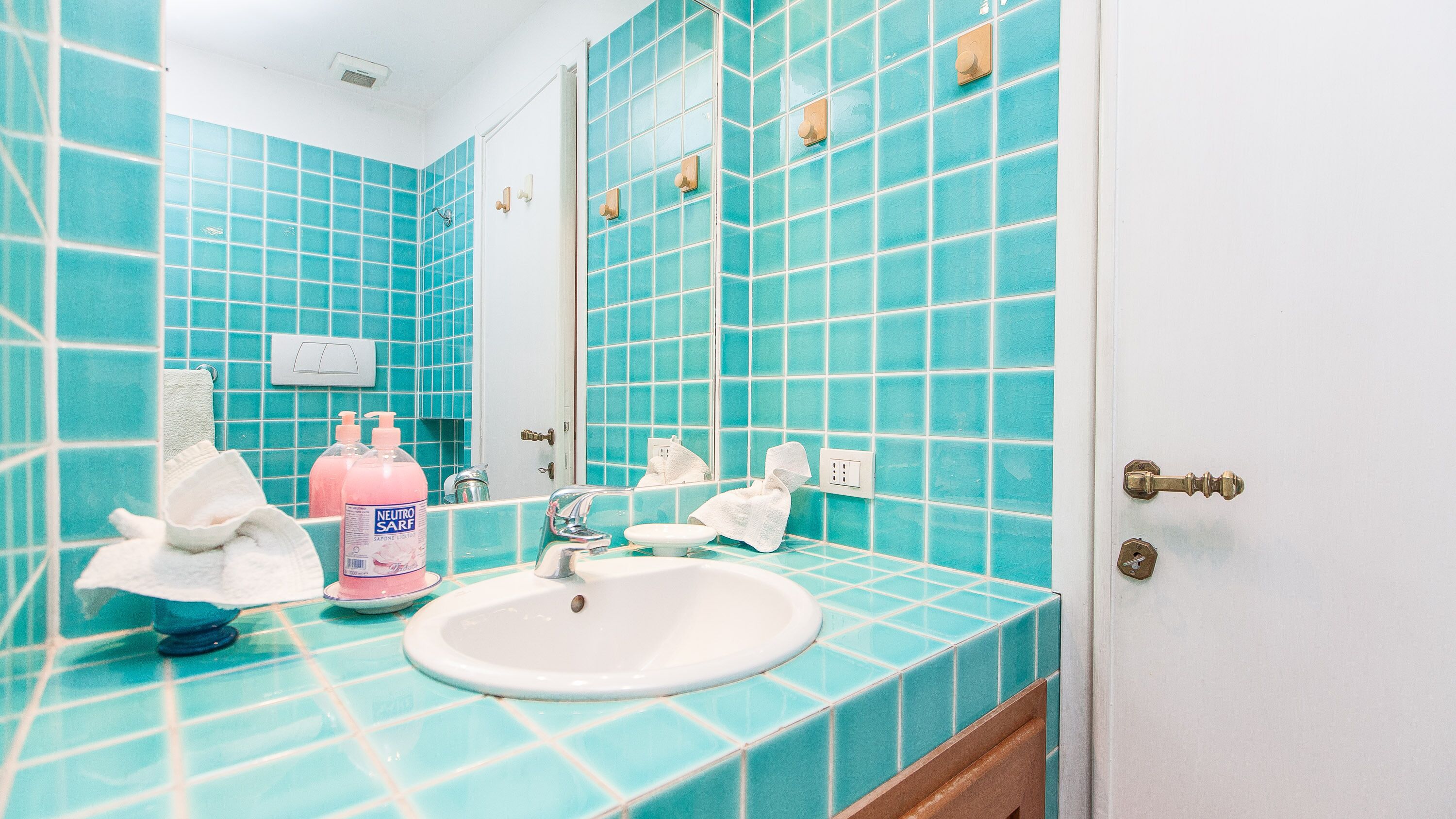 A bathroom with turquoise tiles, a sink, two mirrors, and toiletries.