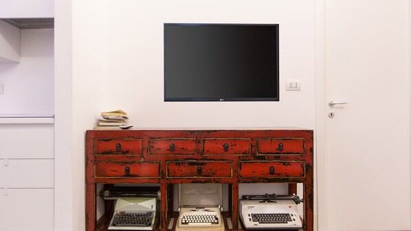 A room with a white wall, a black flat-screen TV mounted above a red wooden cabinet with drawers, and three vintage typewriters on the lower shelf. - Arenula Apartment close to Piazza Navona (Rome)