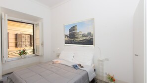 A bedroom with a bed, a window with shutters, a framed picture of the Colosseum, and a small table with flowers. - Arenula Apartment close to Piazza Navona (Rome)