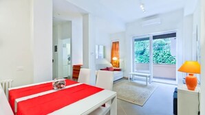 Ein modernes Hotelzimmer mit einer rot-weißen Bettdecke, einem kleinen Tisch mit einem Glasbehälter, einem weißen Sofa, einem großen Fenster mit Blick auf Grünflächen und einer leuchtend orangefarbenen Lampe.