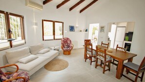 A living room with a white sofa, a wicker chair, a dining table with chairs, and a kitchen area with a refrigerator. - Mimosa - pretty holiday property with private pool in Moraira (Benissa)