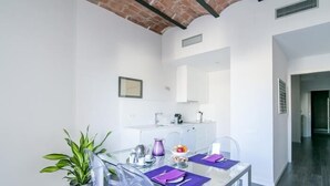 A modern dining area with a glass table, clear plastic chairs, a potted plant, and a kitchen in the background. - One bedroom apartment (Barcelona)