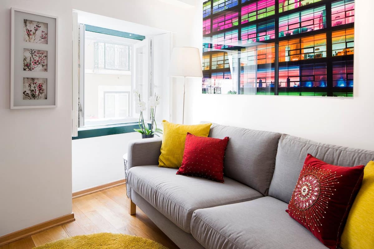 A modern living room with a grey sofa, yellow and red cushions, a white lamp, and a colorful glass wall.
