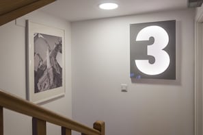 A hallway with a framed abstract painting, a large illuminated number '3', and a wooden staircase railing. - Contemporary by Apartments Alfama (Lisbon)