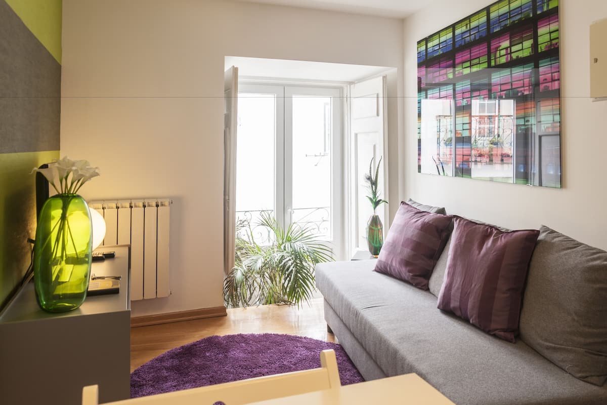 A modern living room with a grey sofa, a purple rug, a green vase with flowers, and a large window with a view of a balcony.