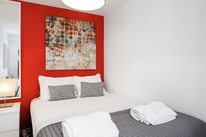 Ein Hotelzimmer mit Bett, einer roten Akzentwand, einem großen abstrakten Gemälde und einem ordentlich bezogenen Bett mit weißen und grauen Textilien.