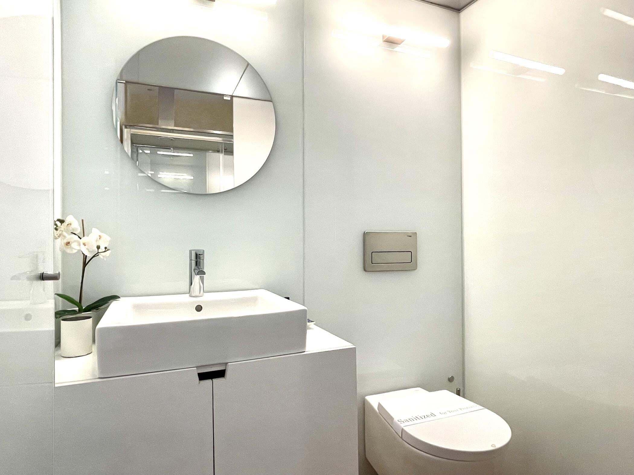 Une salle de bain moderne avec un lavabo blanc, un miroir rond, des toilettes et une petite orchidée en pot.