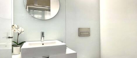 Une salle de bain moderne avec un lavabo blanc, un miroir rond, des toilettes et une petite orchidée en pot.