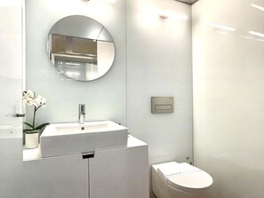A modern bathroom with a white sink, a round mirror, a toilet, and a small potted orchid.