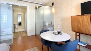 Un espace de vie moderne avec une table de salle à manger ronde, des chaises bleues, un téléviseur posé sur un meuble en bois et une porte en verre donnant sur une salle de bain.