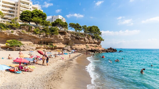 Una playa con gente tomando el sol y nadando, un acantilado rocoso con  árboles, y un edificio al fondo.