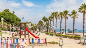 Un parque infantil junto a la playa, con un tobogán rojo y estructuras para escalar, rodeado de palmeras y un cielo despejado.