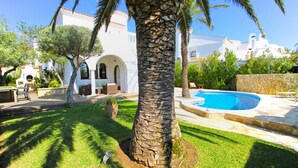 Zona de piscina con una palmera, un muro de piedra y un edificio blanco con arcos al fondo.