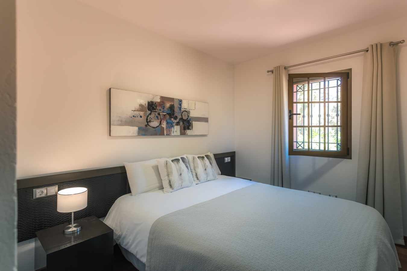 Een keurig opgemaakt hotelkamer met een bed, een bedlampje, een raam met gordijnen en een aan de muur hangend kunstwerk.