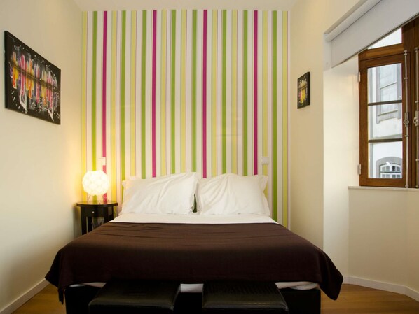 A bedroom with a bed, a bedside table with a lamp, a window, and a striped wall.