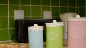 Trois boîtes de rangement pour la cuisine, dotées de couvercles et étiquetées « Thé », « Sucre » et « Café », sont posées sur une surface en bois, avec un pare-feu en carreaux verts derrière.