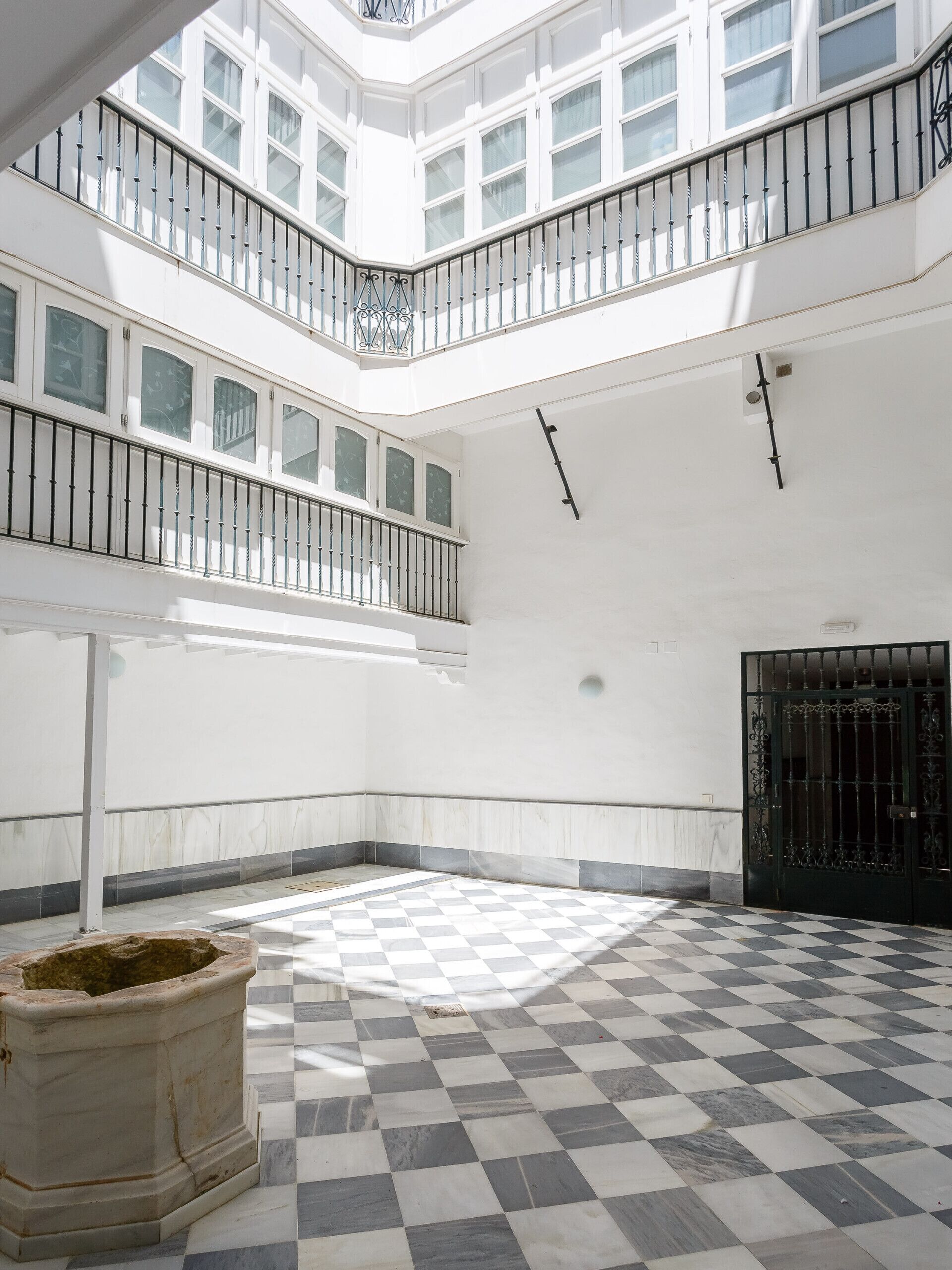 A spacious atrium with a checkered floor, a black gate, and a white wall with windows.