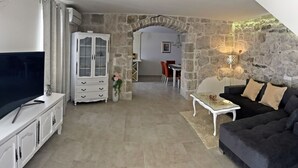 A living room with a TV, white cabinet, black sofa, stone wall, and a dining area in the background.