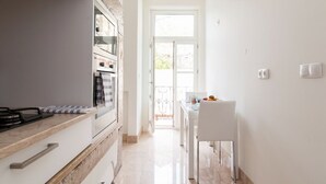 Una cocina con estufa, horno y armarios blancos. Hay una zona de comedor con mesa y sillas, y una puerta que da al exterior.