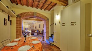 Un comedor con un arco de ladrillo que conduce a una sala de estar. Hay una mesa preparada para cuatro, con platos, vasos y cubiertos. La habitación tiene un techo de madera y una escalera que sube a un nivel superior.