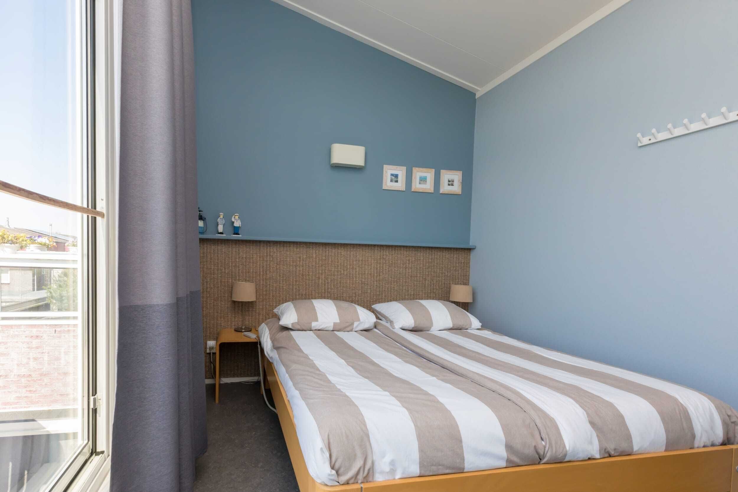 A bedroom with a bed, a nightstand, a window with curtains, and a wall-mounted shelf with items.