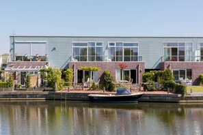 A modern building with large windows, a boat docked in front, and a clear sky. - Complete, popular island house with steam shower. There is a lockable fence between the garden and t (OUDE-TONGE)