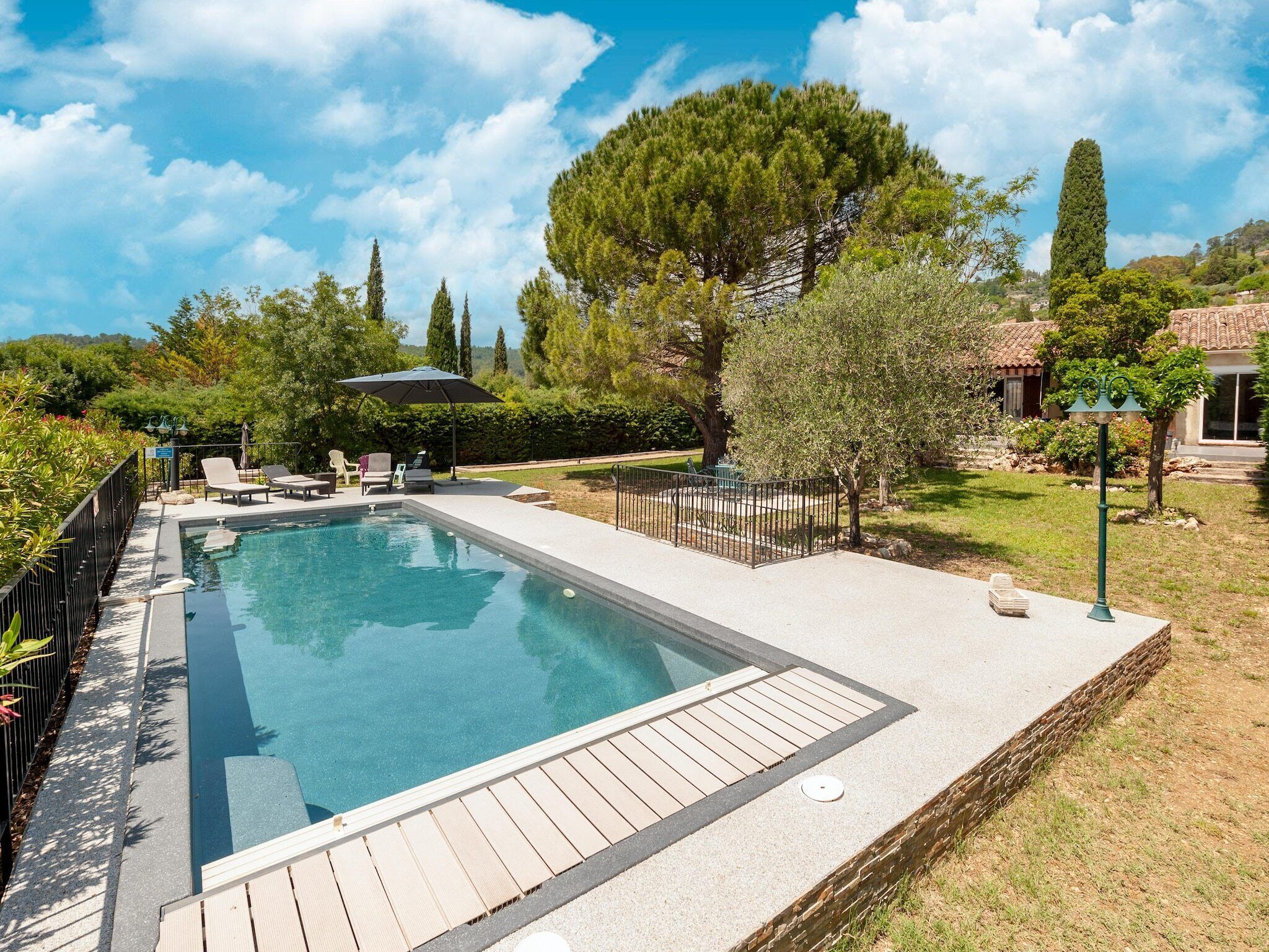 A rectangular swimming pool with a wooden deck, surrounded by a fence, and shaded by a large umbrella.