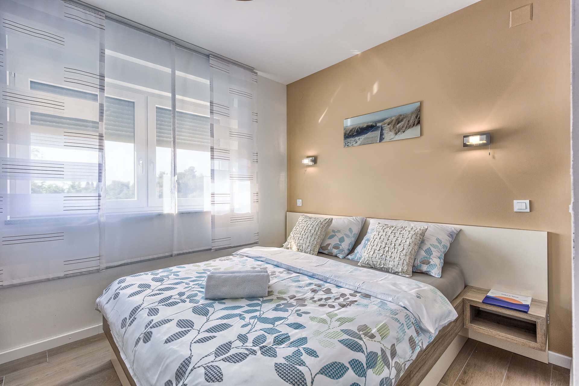 A bedroom with a bed, a window with blinds, a wall-mounted light, and a small bedside table with a book.
