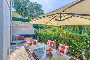 Een terras buiten met een eettafel, omringd door stoelen met geruit kussens, en een grote parasol voor schaduw.