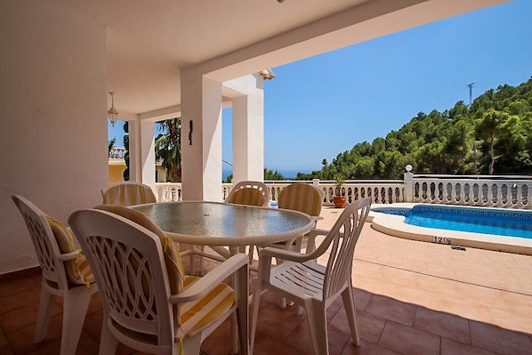 A covered outdoor area with a round table and chairs, a small pool, and a view of a forested hill.