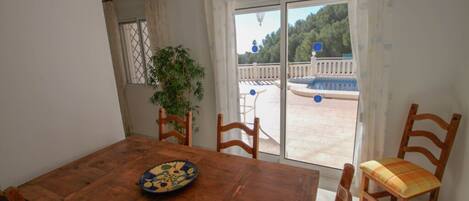 A wooden dining table with chairs, a potted plant, and a view of an outdoor pool and deck through a glass door.