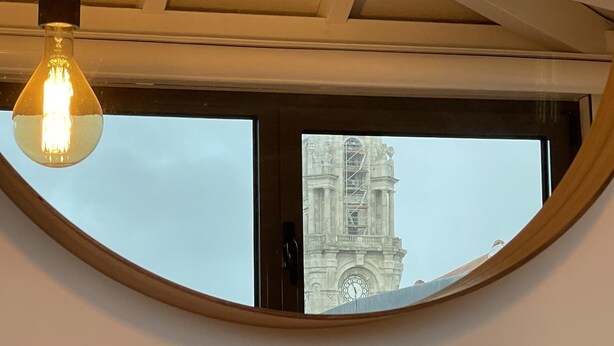A round window with a view of a clock tower through a building's window. - The Brewmaster's House 5 | Rooftop Bedroom (Porto)