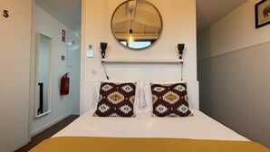 A hotel room with a large bed, two patterned pillows, a round mirror, a wall-mounted light, and a fire extinguisher. - The Brewmaster's House 5 | Rooftop Bedroom (Porto)