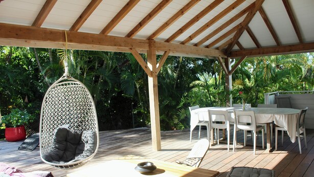 Une terrasse en bois avec un fauteuil suspendu, une table avec des chaises et un espace couvert comprenant une table et des chaises.