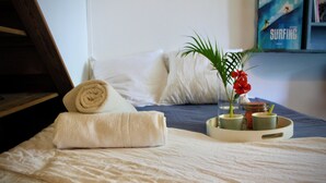 Un lit bien fait, avec une serviette roulée, un plateau avec des tasses et une plante, et un livre intitulé « Surf » accroché au mur.