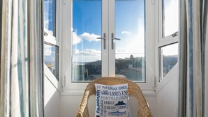 Una silla de mimbre con un cojín que muestra nombres de pueblos costeros, ubicada junto a una ventana con vista despejada del cielo y edificios.