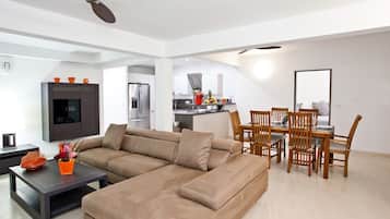 A modern living room with a sectional sofa, a dining area with wooden chairs, a kitchen with a refrigerator and stove, and a ceiling fan.