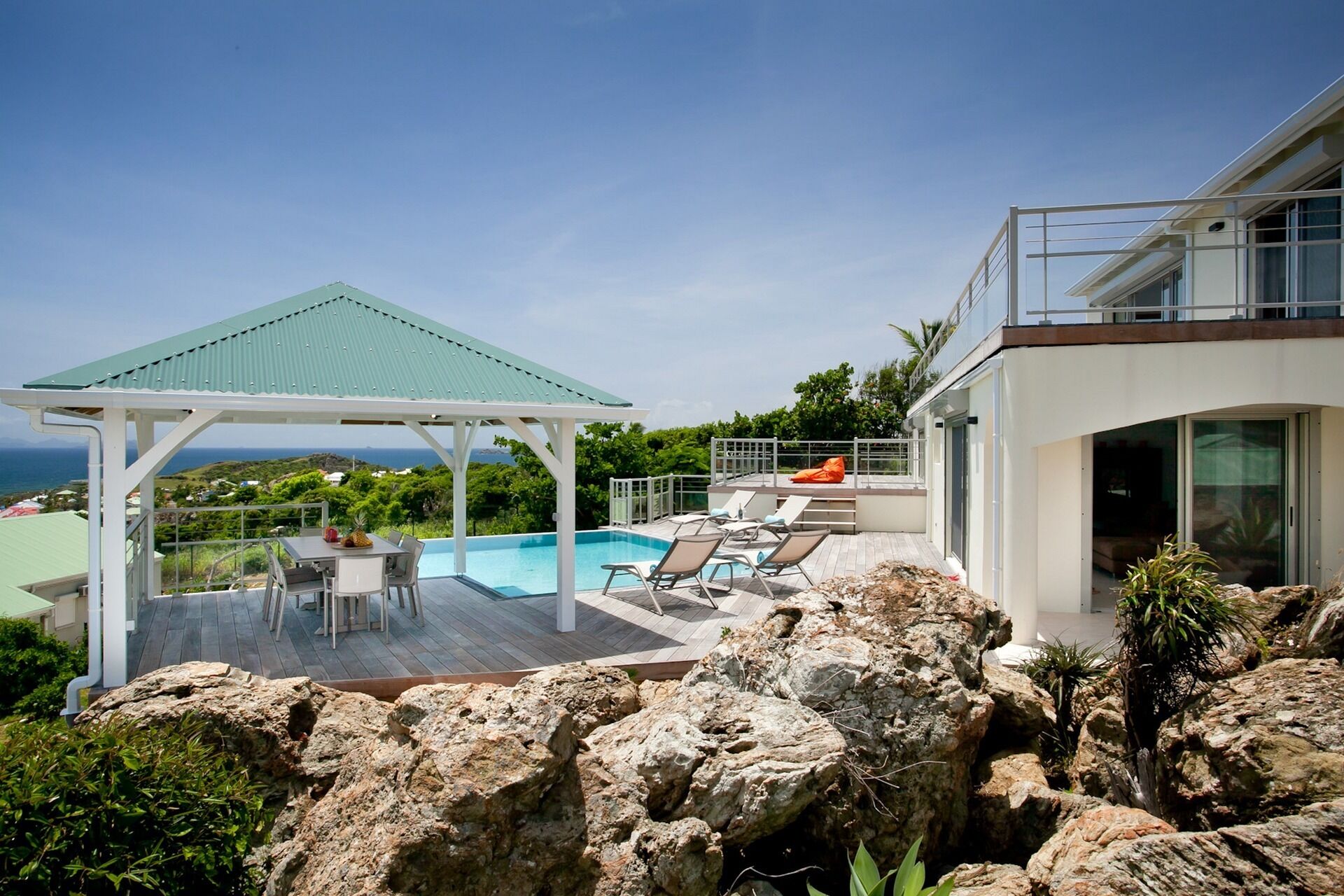 A modern outdoor pool area with a covered seating area, lounge chairs, and a view of the ocean.