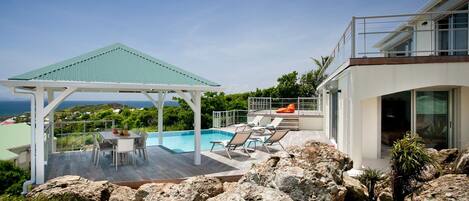 A modern outdoor pool area with a covered seating area, lounge chairs, and a view of the ocean.