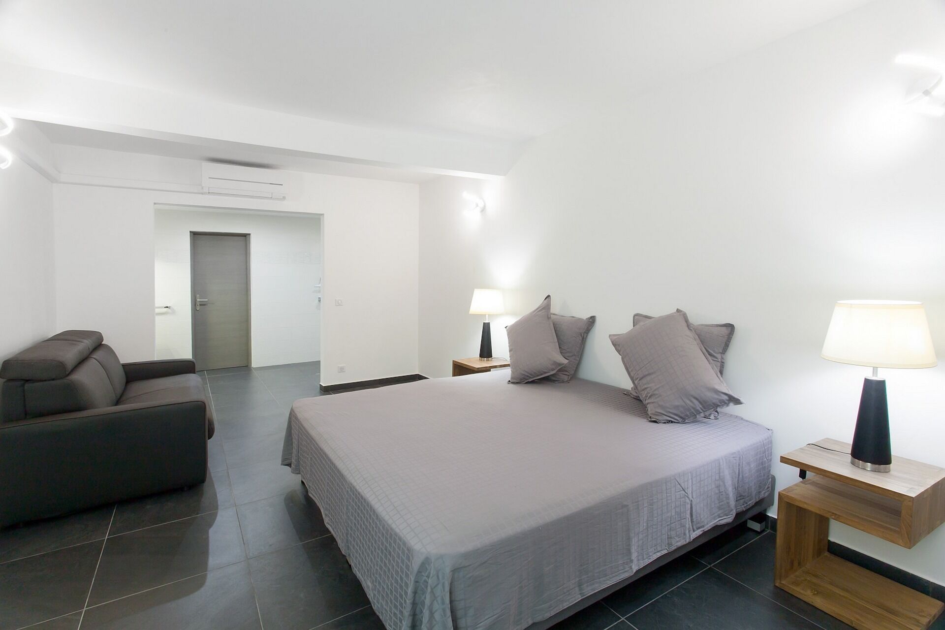 A modern hotel room with a large bed, two bedside tables with lamps, a grey sofa, and a door leading to another room.