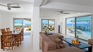 A modern living room with a large sofa, dining area with wooden chairs, and a table with fruit. Large windows offer ocean views. - Le Bougainvillier, magnificent villa with breathtaking views (Oyster Pond)