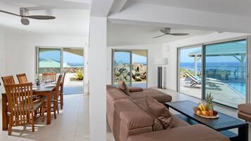 A modern living room with a large sofa, dining area with wooden chairs, and a table with fruit. Large windows offer ocean views.