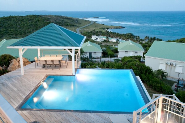 A pool with a view of the ocean, a covered patio, and several buildings with green roofs. - Le Bougainvillier, magnificent villa with breathtaking views (Oyster Pond)
